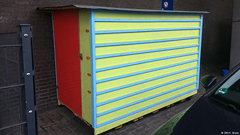 Box in yellow, blue and red, sits on wheels and is lockable home for homeless people. 