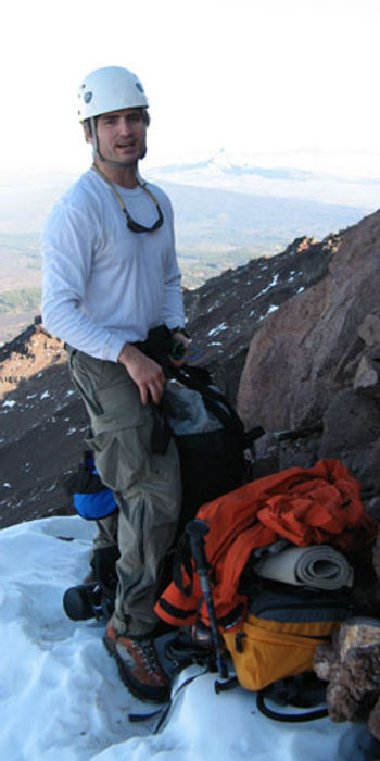 Scott light sitting on his pack during a rest stop on the South Sister in Central Oregon. In the background are other mountains in the Cascade Range and a view of the valley below.