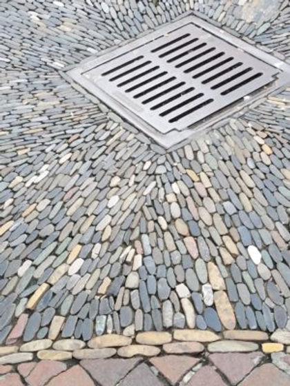 A rush of river rock has a vortex of water going down the storm drain. All in carefully laid out diagonal lines/Richen