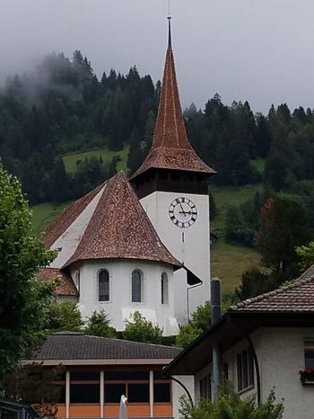 The church is white with brown roofing. It's tower sports a clock and a conical cap-like roof. 